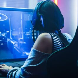 Femeie care se joacă un joc video pe PC, pe ecran sunt orme în nuanțe albastre și mov, ea stă pe scaunul negru, cu căști pe urechi, unele dintre cele mai scumpe jocuri video