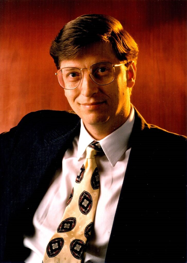 Bill Gates când era tânăr. Poartă un costum negru, cu o cămașă albă, fundal portocaliu