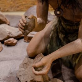Imagine cu om din neanderthal care încearcă să facă focul. poartă blonuri și are pietre in mână