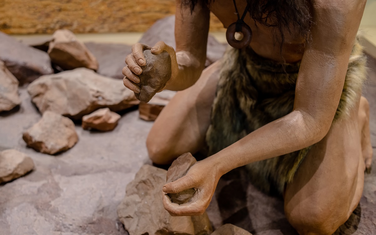 Imagine cu om din neanderthal care încearcă să facă focul. poartă blonuri și are pietre in mână