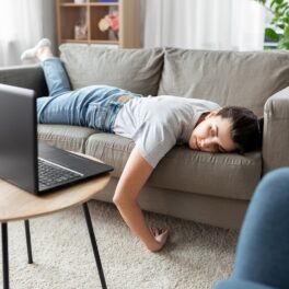 Femeie care stă tolănită pe canapea, cu laptopul pe măsuța mică de lemn din față. Canapeaua e gri. Obiceiul face parte din stilul de viață din pandemie