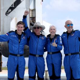 Jeff Bezos și echipa spațială Blue Origin, în anul 2021, toți poartă uniforme albastre. au o navetă spațială în spate