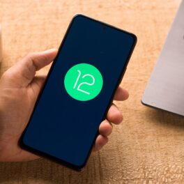 Telefon negru, cu ecran negru, care are logo-ul verde Android 12 pe ecran. Telefonul e ținut în mână pe un birou din lemn, cu un laptop lângă Android 12 introduce noi opțiuni