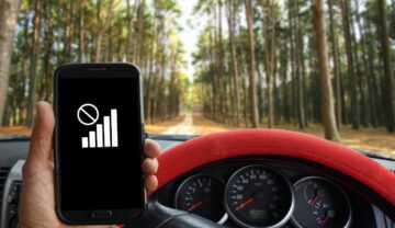 Utilizator care ține un smartphone în mână fără semnal, la un volan roșu, cu pădure în față, se văd barele de semnal de pe telefon