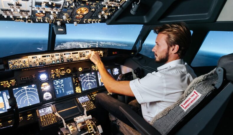 Pilot blond, în cabina de zbor a unui avion, cu multe butoane în față, stă pe scaun, geamurile închise, ocean și un oraș pe fundal. Acest pilot nu deschide geamul avionului în timpul zborului