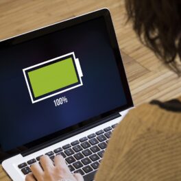 Laptop cu baterie încărcată la maxim, baterie verde pe ecran negru, cu o femeie în fața lui. Mulți se întreabă ce se întâmplă dacă încarci laptopul toată noaptea