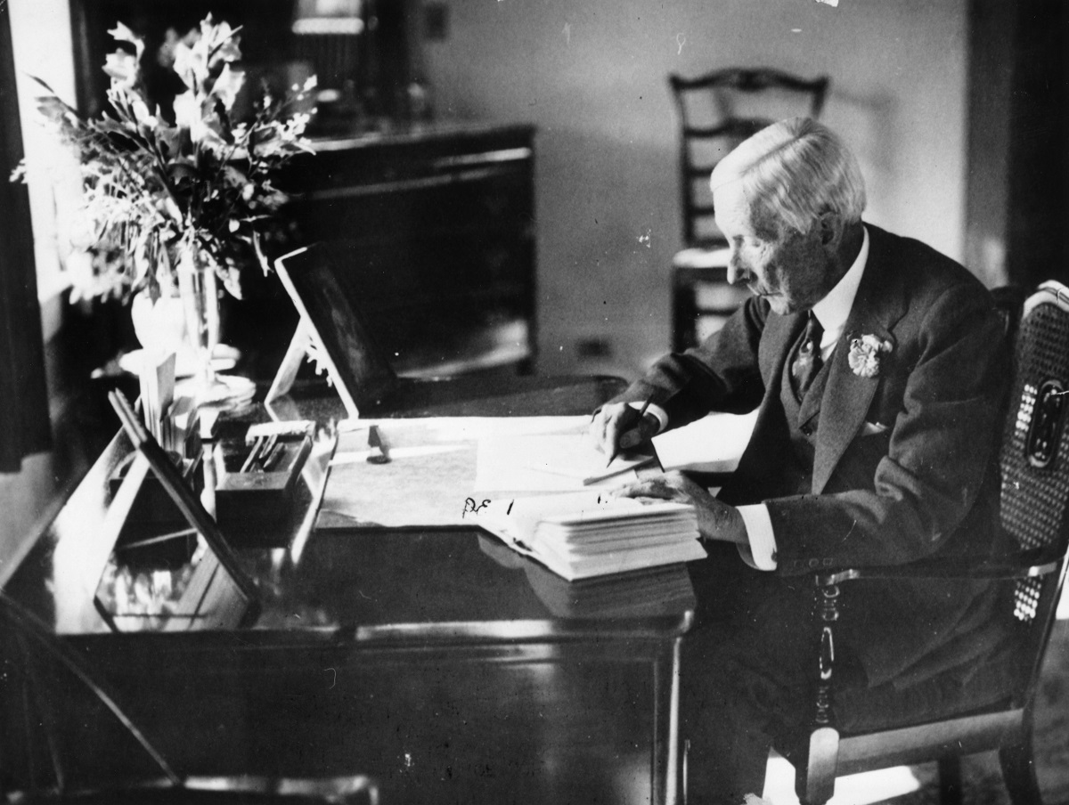 John D. Rockefeller stă în scaun, la birou, și scrie. E îmbrăcat în costum. Fotografie alb-negru. El a devenit primul miliardar din istoria SUA