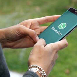 Persoană care ține în mână un smartphone negru, cu logo-ul WhatsApp pe ecranul verde. Fundal cu verdeață. Mulți utilizatori nu știu cum să verifice dacă ai fost blocat pe WhatsApp