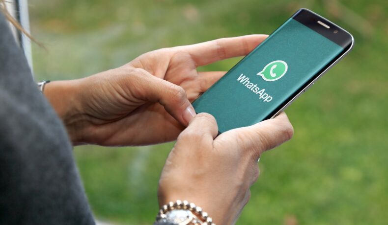 Persoană care ține în mână un smartphone negru, cu logo-ul WhatsApp pe ecranul verde. Fundal cu verdeață. Mulți utilizatori nu știu cum să verifice dacă ai fost blocat pe WhatsApp