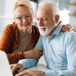 Un bărbat și o femeie în vârstă care se uită la un laptop gri deschis. Amândoi stau jos i se uită la ecran. Bărbatul are o cămașă albastră, femeia e îmbrăcată în maro. Un posibil secret al longevității centenarilor se află în sistemul digestiv