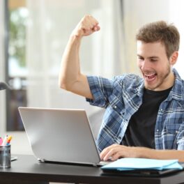 Bărbat în fața unui laptop, joacă jocuri video, mâna în sus. Laptopul e gri. Experții au analizat cum pot schimba jocurile video comportamentul jucătorilor