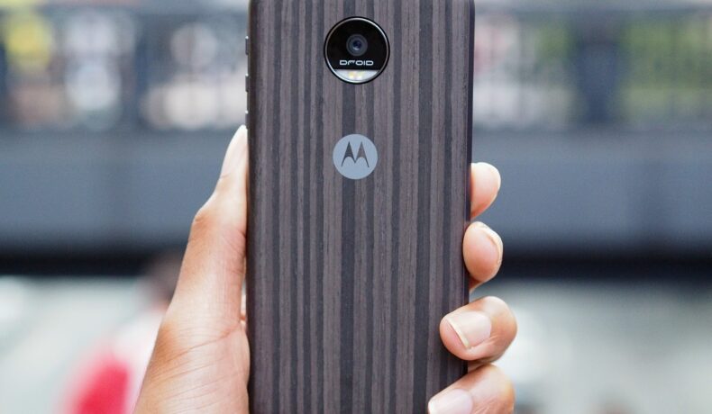 Moto Z, fotografiat în 2016. Telefonul mobil are carcasă cu maro, negru și gri, spate, cu logo-ul Motorola, ținut în mână. Nu seamănă cu Moto G200 5G