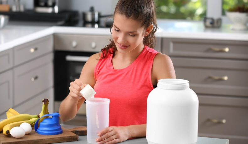 Femeie care stă în bucătărie și își prepară shake de proteine, pe fundal gri. Provocarea virală de pe Tiktok include pudra de proteine pre-antrenamet