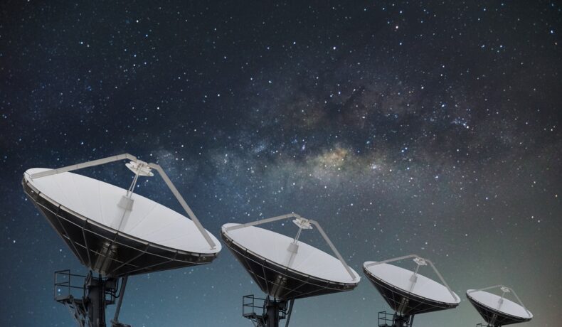 Cercetatorii cred că au aflat ce cauzează semnalul misterios din Calea Lactee. În imagine sunt trei radare mari, albe, cu cerul cu calea lactee pe fundal
