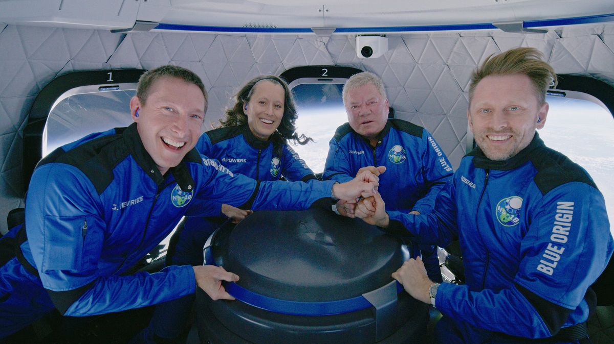 William Shatner și echipajul navetei Blue Origin care a ajuns în spațiu în octombrie 2021. Sunt toți îmbrăcați în costume albastre, stau la masă și se țin de mână