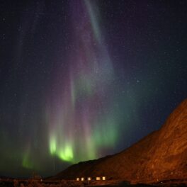 Aurora Boreală văzută pe cerul Groenlandei, în nuanțe de verde și mov, cu un munte în partea dreaptă