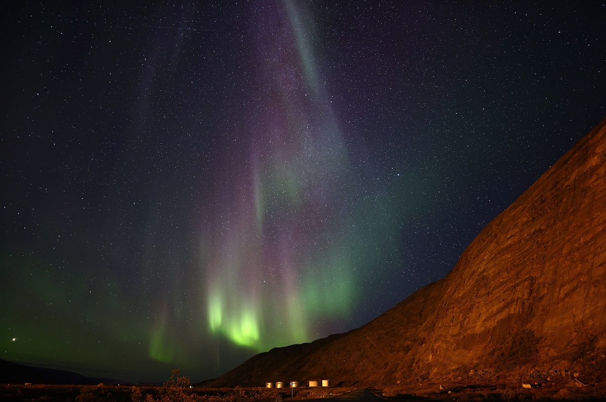 Aurora Boreală văzută pe cerul Groenlandei, în nuanțe de verde și mov, cu un munte în partea dreaptă