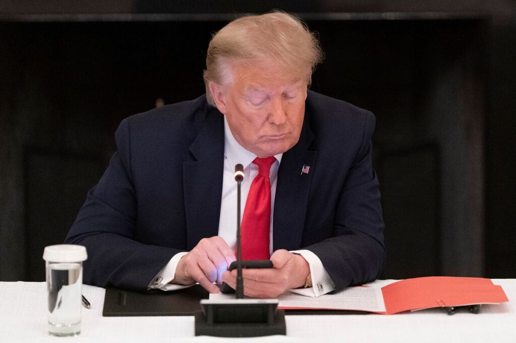 Donald Trump la o conferință de la Casa Albă. Stă la masă, pe scaun, are un smartphone în mână, se uită peste un dosar. Donald Trump va lansa o rețea de socializare în curând