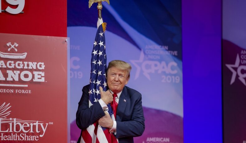 Donald Trump ia în brațe steagul american în cadrul unui eveniment. Fundalul e cu roși și albastru