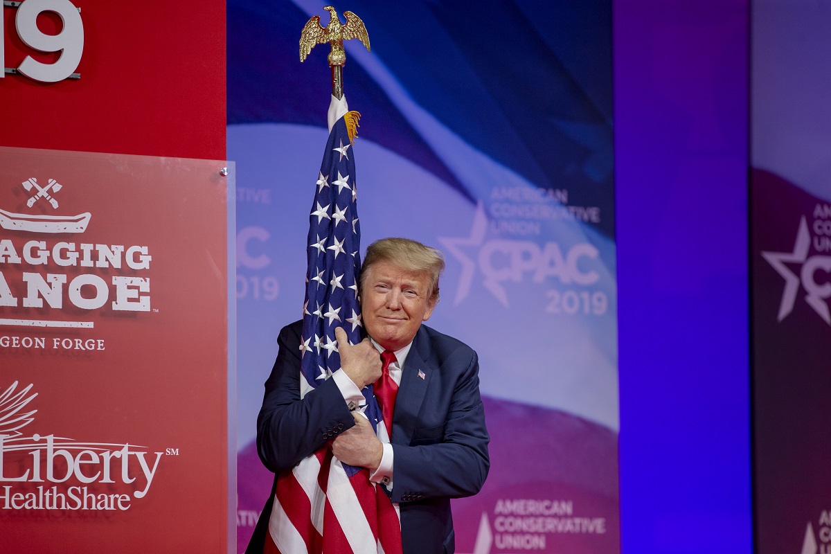 Donald Trump ia în brațe steagul american în cadrul unui eveniment. Fundalul e cu roși și albastru