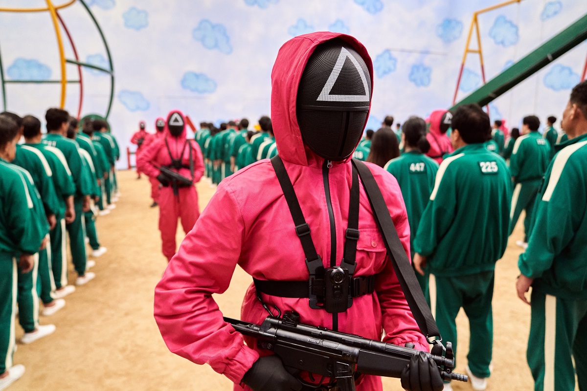 Gardien din serialul Squid Game, îmbrăcat în roz, care merge printre rândurile de concurenți, cu o armă