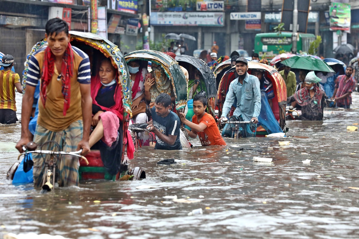 Inundație în Bangladesh, cu un șir de oameni care trec prin apa care le vine până la genunchi. Sunt îmbrăcați în haine colorate, primul bărbat merge pe o bicicletă