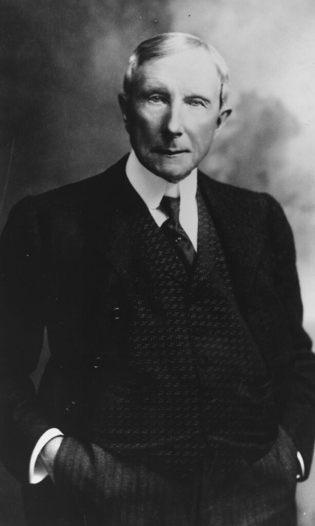 John D. Rockefeller, bătrân. Poartă un costum negru, cămașă albă și cravată