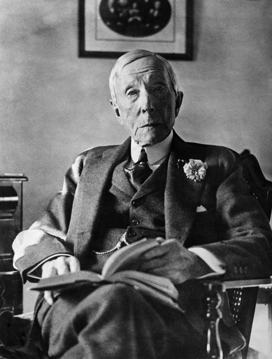 John D. Rockefeller, în ultimii ani din viață, stând pe un scaun. E îmbrăcat în costum închis la culoare, cu cămașă și cravată. Are o carte deschisă în poală