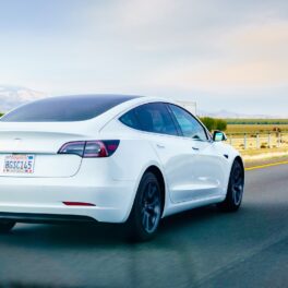 Mașină Tesla Model 3, varianta pe alb, care merge pe o șosea rurală, cu peisaj verde și cer albastru pe fundal