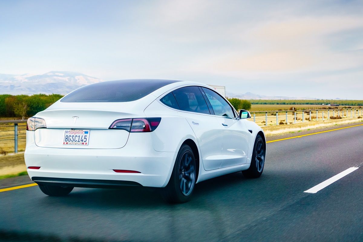 Mașină Tesla Model 3, varianta pe alb, care merge pe o șosea rurală, cu peisaj verde și cer albastru pe fundal
