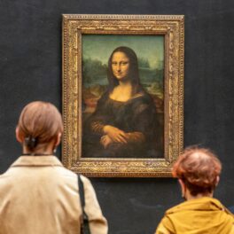 Mona LIsa (La Gioconda), creată de Leonardo da Vinci, expusă în muzeul Louvre. Peretele e albastru, rama e aurie, două persoane stau și se uită la ea