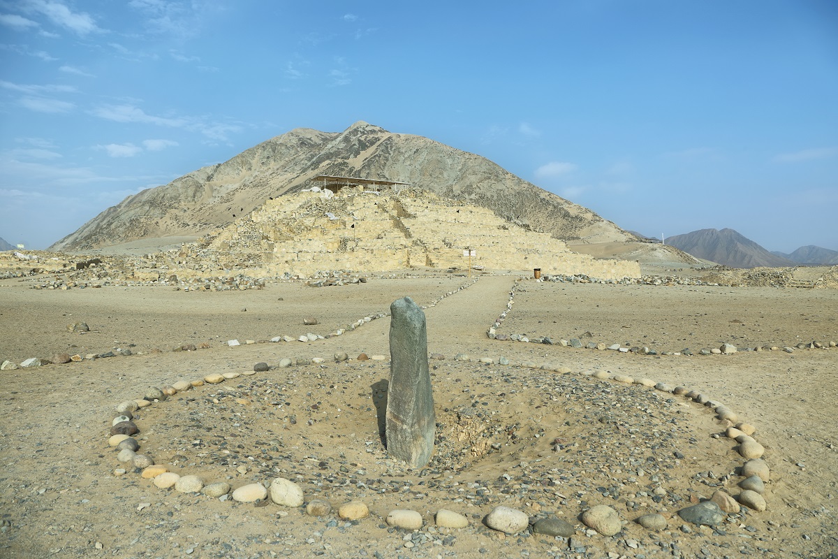 Orașul Caral, din Peru, include undele dintre cele mai vechi piramide din lume. Semn de piatră în centru, cu piramida pe fundal. Cerul e albastru