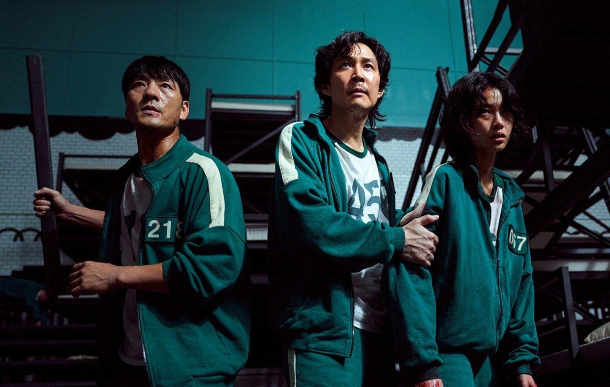 Lee Jung-jae și alți doi actori, îmbrăcați în trening verde toți 3. Sunt înarmați și stau într-o cameră întunecată