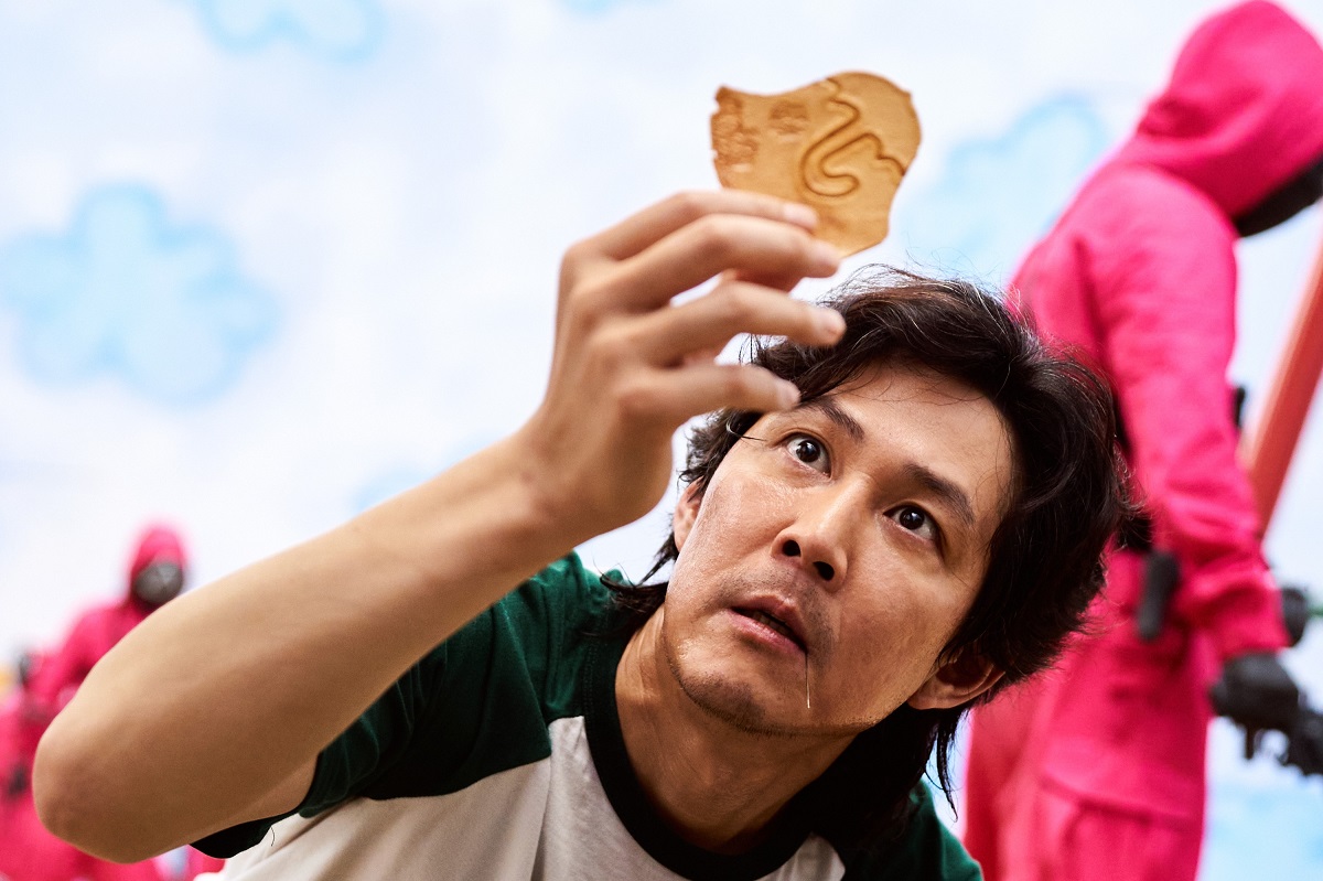 Lee Jung-jae în serialul Squid Game, ce a avut un profit uriaș. Ține o prăjitură în mână, are un tricou cu alb și verde, cer albastru pe fundal
