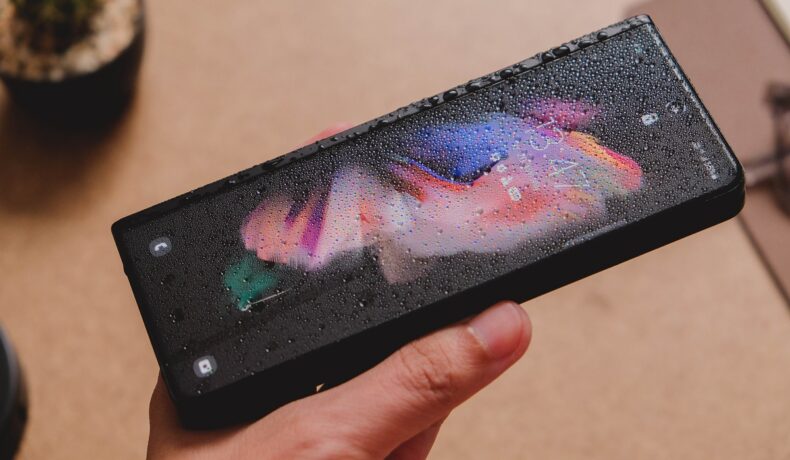 Telefon Samsung Galaxy Z Fold 3, ținut în mână, cu picături de apă pe ecran. E pliat, fundal de lemn