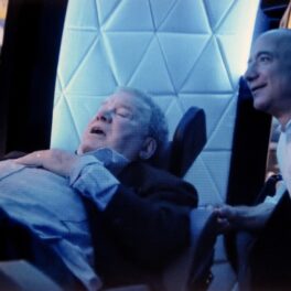 William Shatner și Jeff Bezos vorbesc. Shatner se află într-un fotoliu mare și alb, Jeff Bezos e lângă el. Shatner poartă un costum închis la culoare, Bezos poartă o cămașă albă