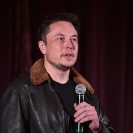 Elon Musk la o conferință tech din anul 2018. Poartă o geacă de piele maro, cu guler bej, roșu pe fundal. Acțiunile Tesla au scăzut puternic recent