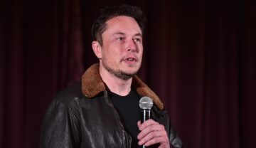 Elon Musk la o conferință tech din anul 2018. Poartă o geacă de piele maro, cu guler bej, roșu pe fundal. Acțiunile Tesla au scăzut puternic recent