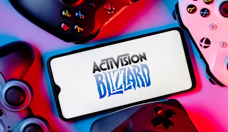 Logo-ul Activision Blizzard pe ecranul unui smartphone, înconjurat de controllere pentru console, cu o lumină roșie pe fundal. Activision Blizzard ar fi pierdut milioane de jucători