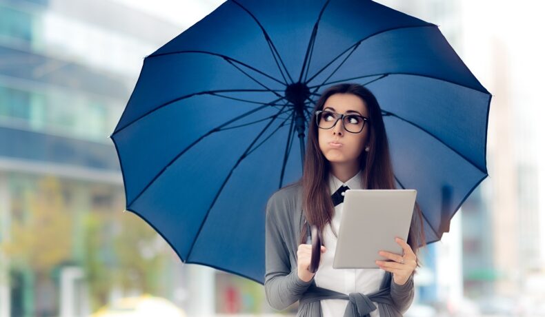 Femeie care stă în ploaie, cu o umbrelă imensă albastră, îmbrăcată într-un costum, cu o tabletă gri în mână. Afară plouă și ea e în oraș. Adevărul despre cum afectează vremea Internetul a fost dezvăluit de experți