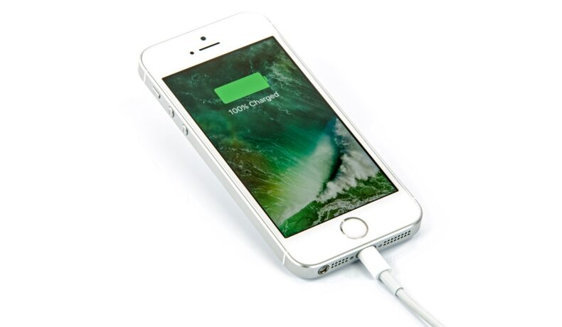Telefon iPhone SE, în imagine oficială Apple, în care se află la încărcat. Telefonul e gri, ecranul e verde și arată procentul bateriei. Mulți se înreabă cum afli procentul bateriei la iPhone 13, dar există un truc simplu