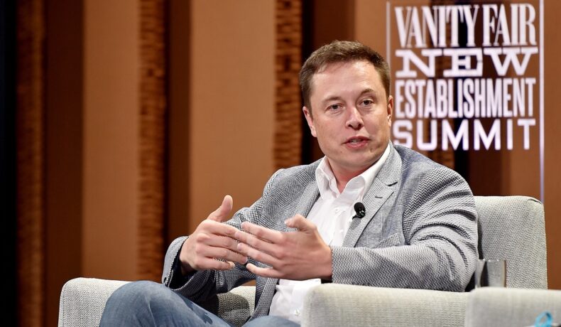 Elon Musk, la o conferință în anul 2015. Poartă un costum gri, blugi, cămașă albă, are un fundal din lemn, stă într-un fotoliu gri. Mulți se întreabă cum e Elon Musk în viaaț reală