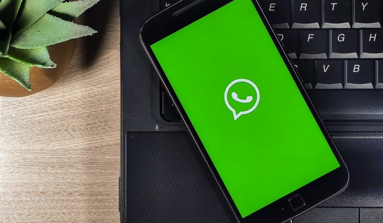 WhatsApp pe ecranul verde al unui telefon, cu o carcasă neagră, care se află pe o tastatură de laptop neagră, pe un birou de lemn. Mulți nu știu cum poți trimite un mesaj WhatsApp