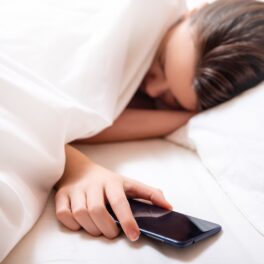 Femeie care doarme în pat, cearșaf alb, care ține un telefon negru în mână. Mulți experți recomandă să nu dormi cu telefonul în pat