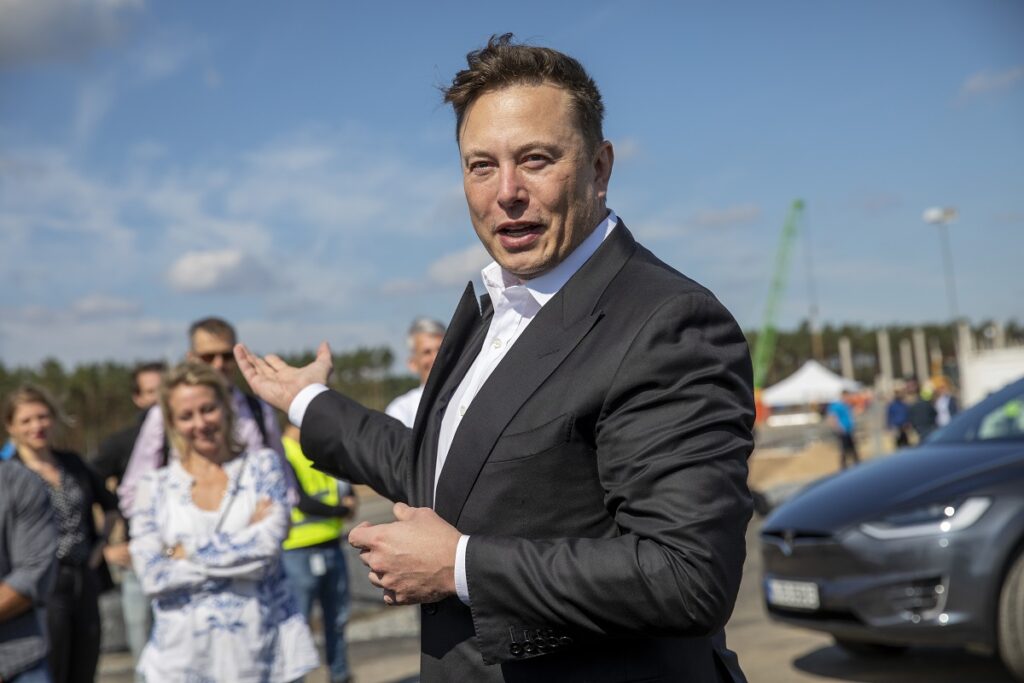 Elon Musk la o conferință de presă din Germania, în anul 2020. Poartă un costum negru, cămașă albă și are cer și mulțime pe fundal. Elon Musk a creat un sondaj pe Twitter recent despre averea sa
