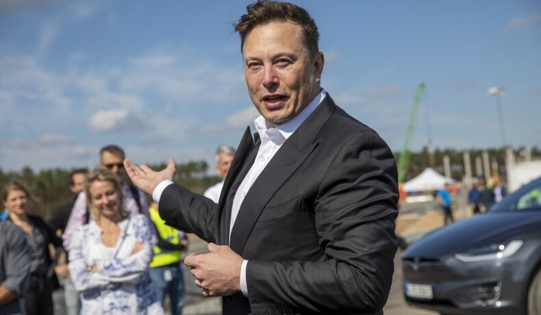 Elon Musk la o conferință de presă din Germania, în anul 2020. Poartă un costum negru, cămașă albă și are cer și mulțime pe fundal. Elon Musk a creat un sondaj pe Twitter recent despre averea sa