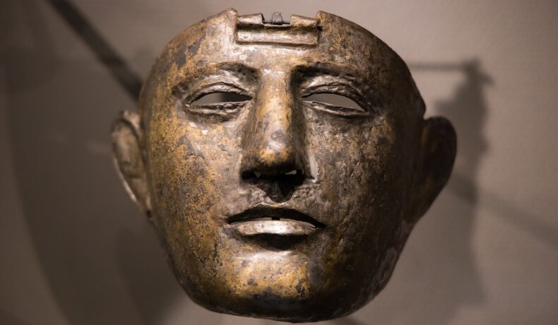 Masca unui soldat roman, expusă în muzeu. E din metal închis la culoare, bej pe fundal. Masca unui soldat roman a fost găsită în Turcia