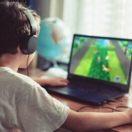 Copil care se joacă un joc video pe un laptop, cu căștile pe cap. Pe laptop, ecranul e verde. Jocul video cu o pisică neagră se numește Little Kitty, Big City