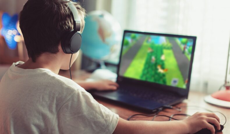 Copil care se joacă un joc video pe un laptop, cu căștile pe cap. Pe laptop, ecranul e verde. Jocul video cu o pisică neagră se numește Little Kitty, Big City