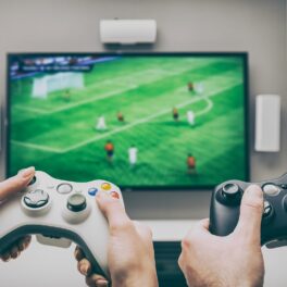 Doi jucători stau în fața unui ecran, pe care e un joc de fotbal. Au controllers în mână, unul alb, celalălalt negru. Numeroase jocuri pentru PlayStation și Xbox sunt îndrăgite în întreaga lume